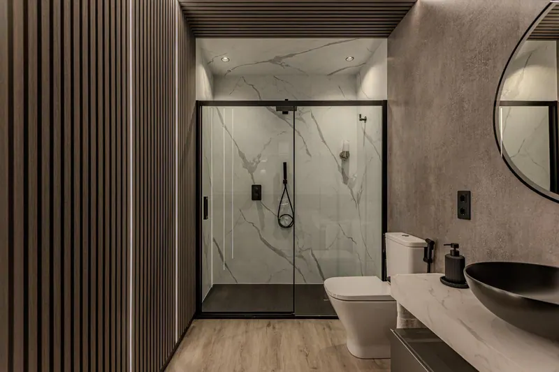 Ducha con paneles grandes de efecto mármol, una pared de lamas de madera y un revestimiento de piedra gris, con luz cálida integrada