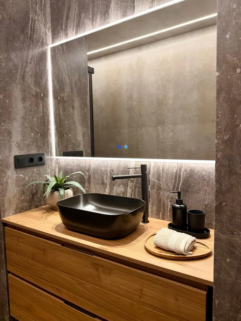 Baño con una pared de revestimiento efecto piedra gris, un mueble de madera con lavabo negro sobre encimera y un espejo retroiluminado