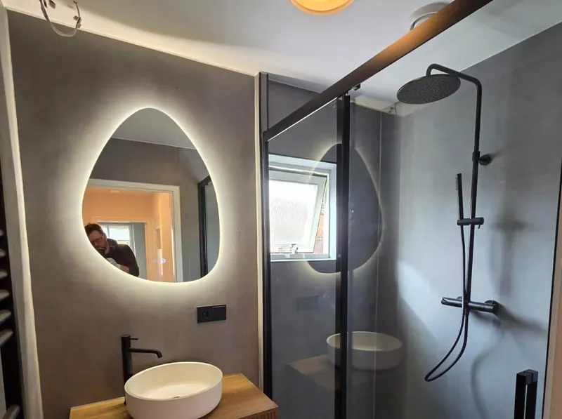 Baño con revestimiento gris en las paredes, un espejo con forma de gota iluminado, un lavabo sobre mueble de madera y una mampara de ducha negra